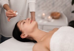 Crop unrecognizable cosmetician applying serum essence on face of young calm female client, lying on couch with closed eyes during beauty procedure in modern salon